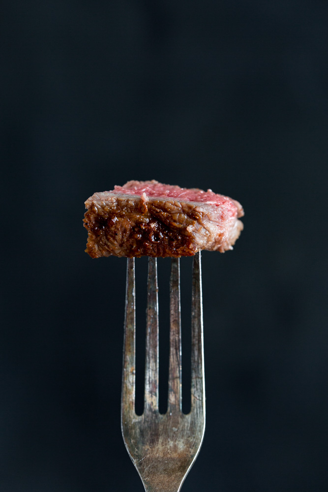 A close-up of a piece of cooked steak with a pink center held by a silver fork against a dark background.