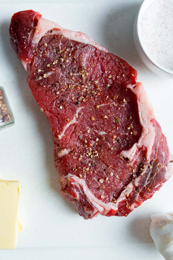 A raw, seasoned beef steak on a white surface next to a bowl of salt, a garlic clove, and a pat of butter.