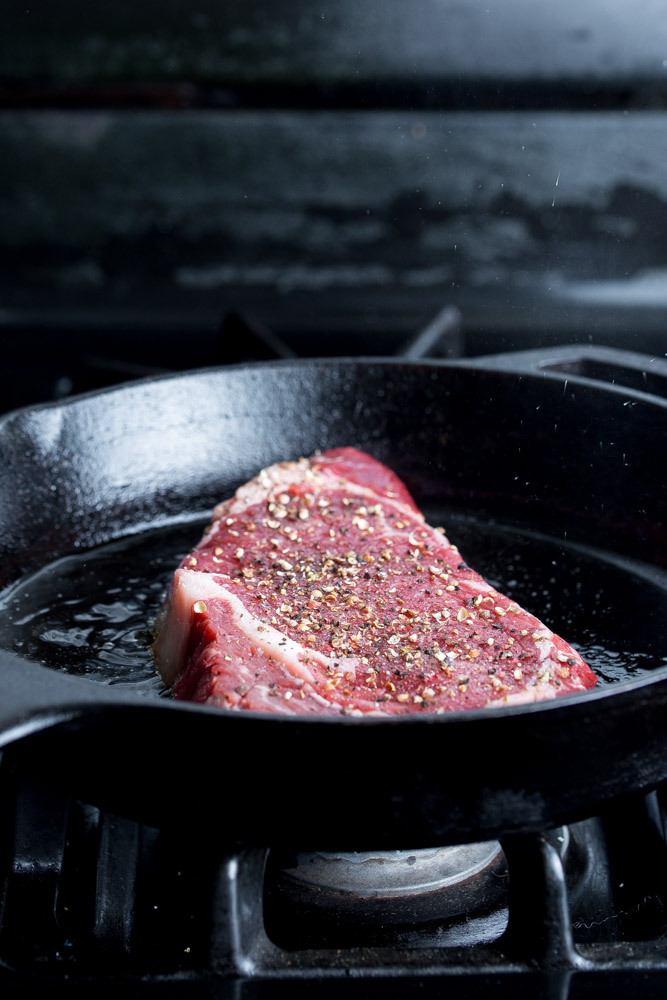 A seasoned piece of raw steak sits in a sizzling black cast-iron skillet on a stove.