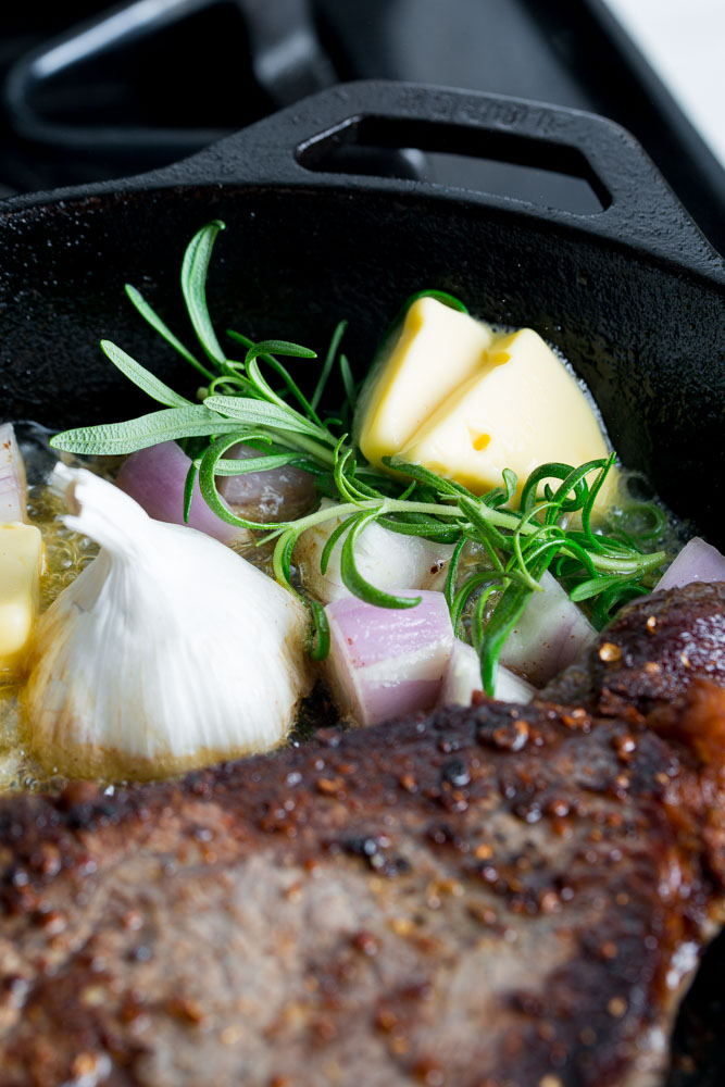 A close-up of a cast iron skillet with a seared steak, rosemary, garlic, shallots, and melting butter.