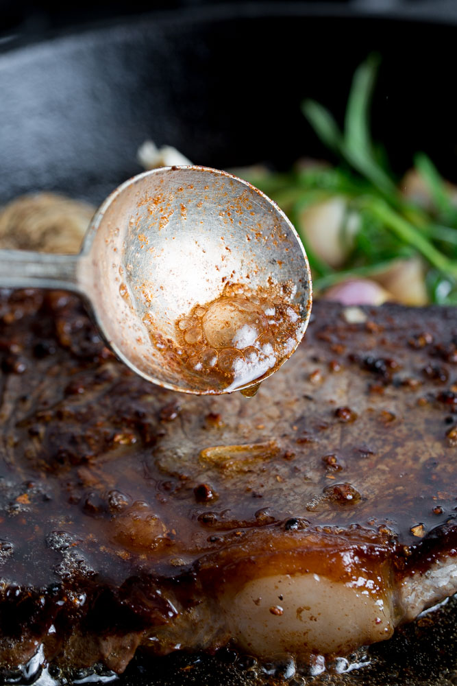 Close-up of a metal spoon pouring sauce over a sizzling steak in a skillet, with greens in the background.