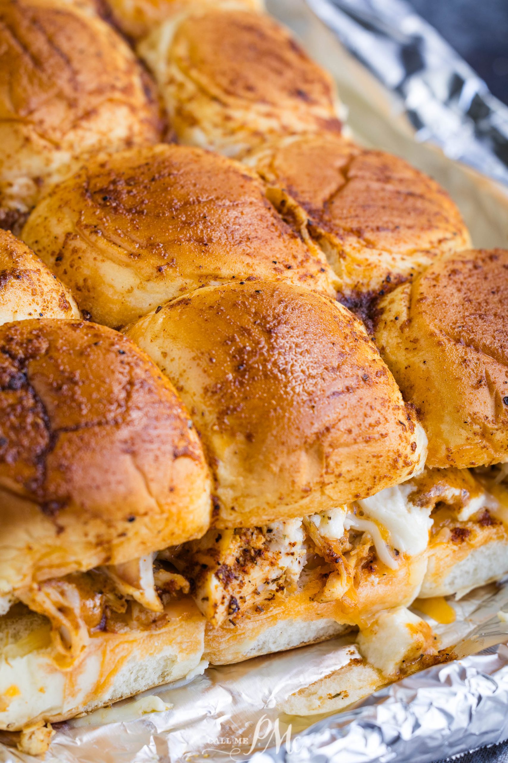 A close-up of a tray of baked sliders, featuring golden brown buns with a crispy top and melted cheese oozing out from between the layers. The tray is lined with aluminum foil.