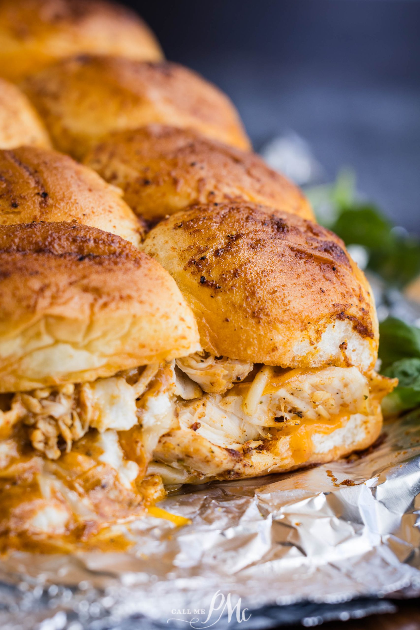 Close-up of seasoned, pulled chicken and melted cheese stuffed inside soft rolls. The sandwiches are placed on aluminum foil.