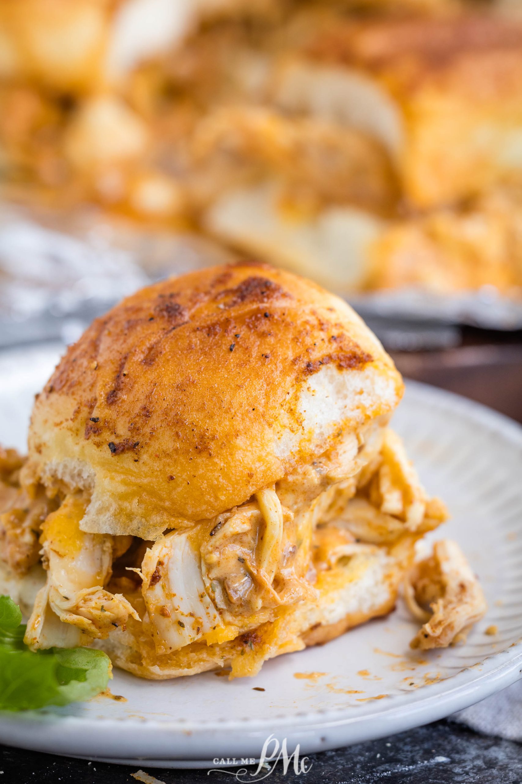 A close-up of a sandwich with shredded chicken and melted cheese on a plate, with more sandwiches and a green garnish in the background.