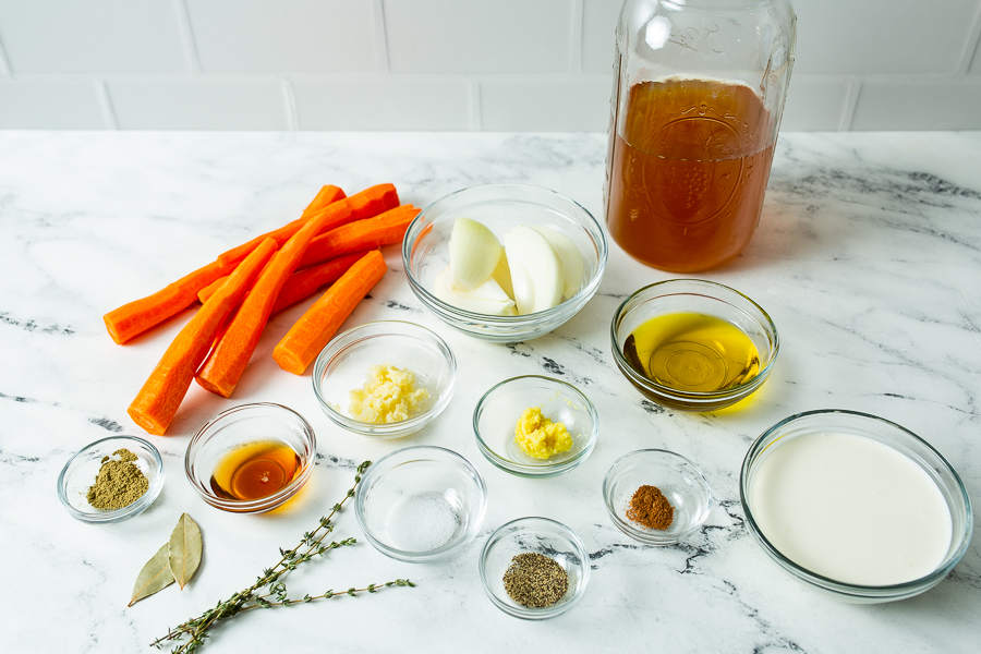 Ingredients assembled on a marble countertop including carrots, onions, garlic, herbs, spices, oil, broth, and cream.