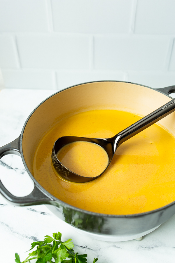 A black ladle rests in a pot of creamy orange soup on a white surface, with a small bunch of parsley nearby.