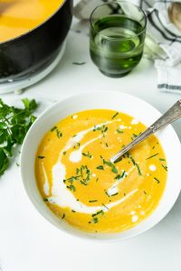 A bowl of creamy orange soup garnished with chopped herbs and a swirl of cream sits on a white table with a spoon, a green glass, and a pot in the background.