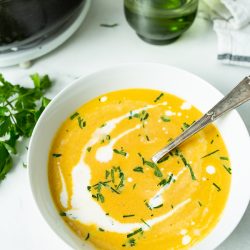 A bowl of creamy orange soup garnished with chopped herbs and a swirl of cream sits on a white table with a spoon, a green glass, and a pot in the background.