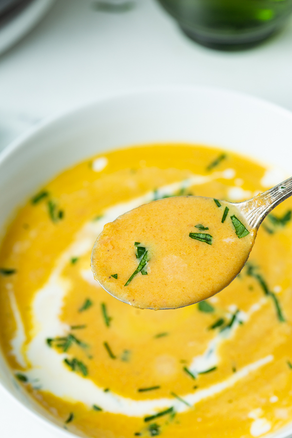 Close-up of a spoonful of creamy soup garnished with chopped herbs, hovering above a bowl filled with the same soup. White drizzle and herbs are visible on the surface of the bowl's contents.