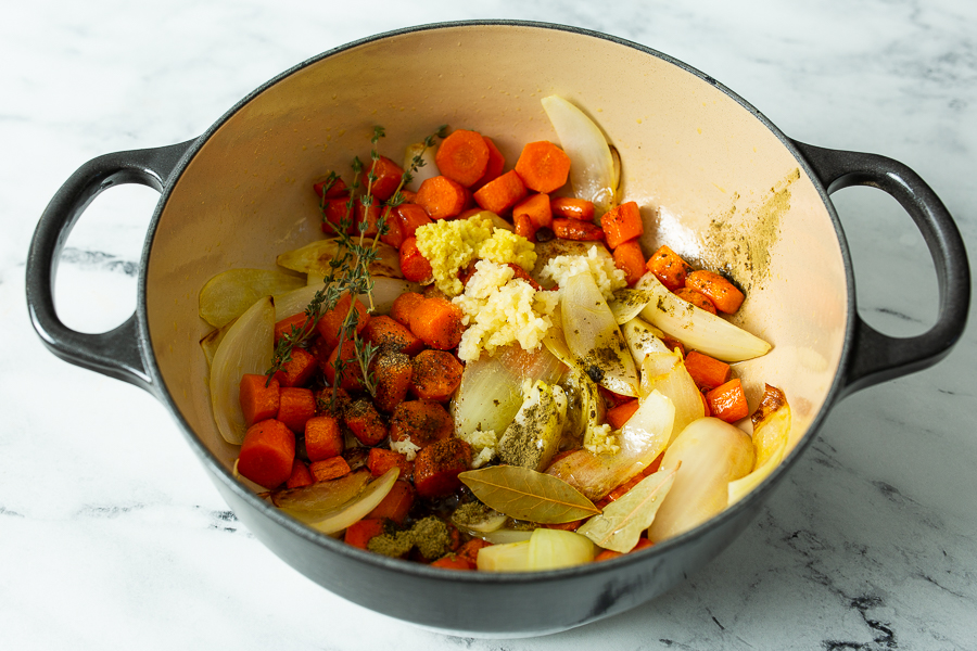 A pot containing chopped carrots, onions, garlic, bay leaves, and herbs, being prepared for cooking on a marble countertop.