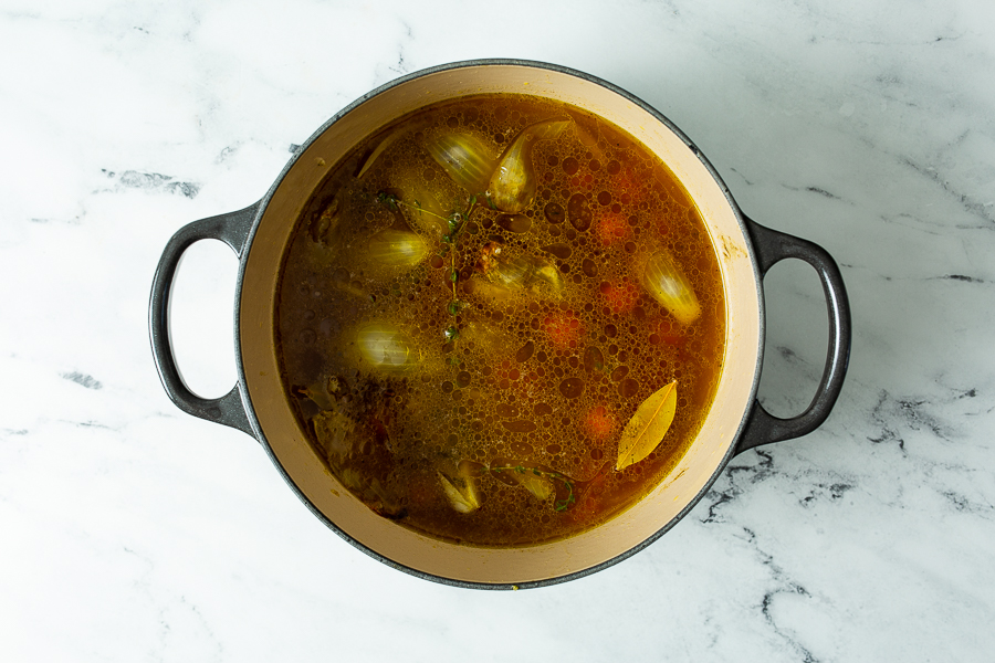 A pot of soup containing whole onions, herbs, and a clear broth sits on a marble countertop.