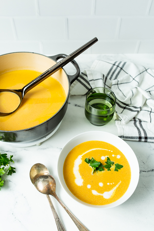 A bowl of Carrot Ginger Soup garnished with parsley, next to a pot of the same soup. Two spoons, a glass of green liquid, and a striped cloth are also on the marble countertop.