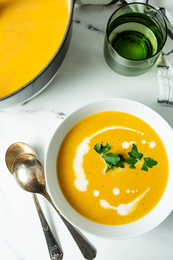 A bowl of Carrot Ginger Soup topped with cream and parsley is next to two silver spoons and a green glass of liquid on a white marble surface.