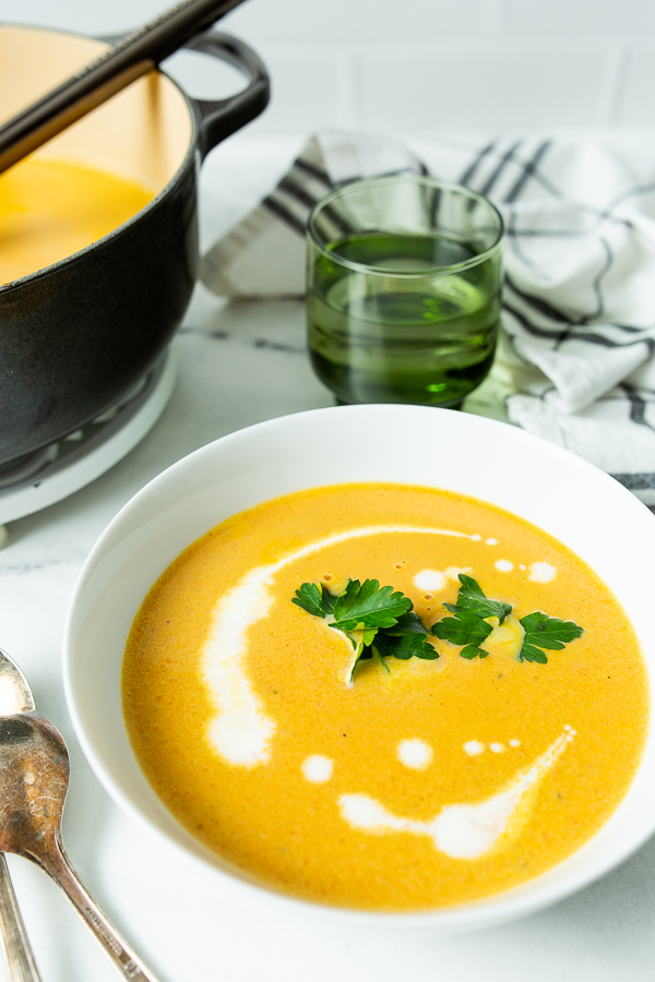 A bowl of Carrot Ginger Soup garnished with parsley and a swirl of cream, next to a spoon, with a pot of soup and a green glass in the background.