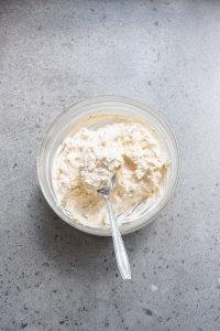 A glass bowl filled with a creamy mixture and a spoon placed inside on a gray surface.