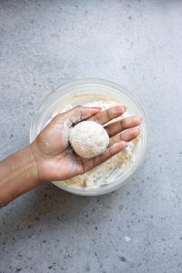 A hand holds a round, dough-covered cheese ball over a bowl filled with more cheese mixture on a gray countertop.