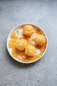 Four round cheese balls coated in a crispy golden crust are placed in a white bowl on a gray surface, with some crumbs scattered around.