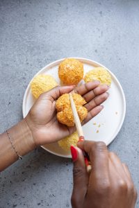 Two hands shaping and pressing a breaded ball of dough over a plate with other breaded balls on a gray surface.