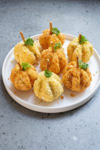 A white plate with seven pumpkin-shaped cheese balls, garnished with pretzel stick stems and green parsley leaves, sits on a gray speckled surface.