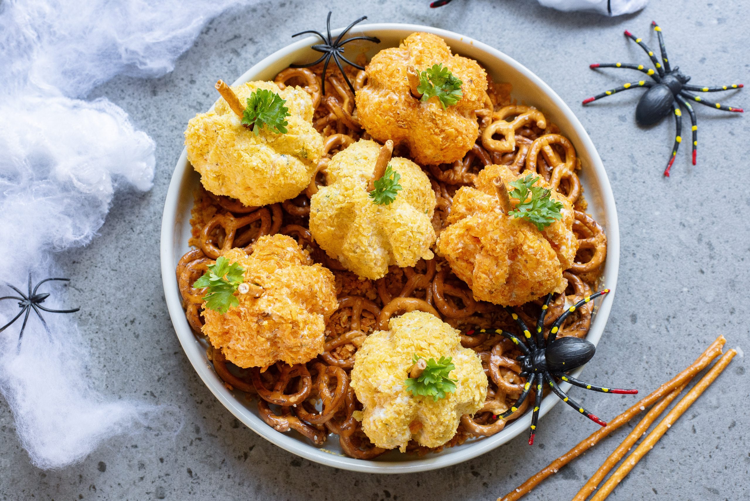 A bowl with Halloween-themed food decorations, featuring cheese balls shaped like pumpkins placed on a bed of pretzels. Plastic spiders and webs surround the bowl.