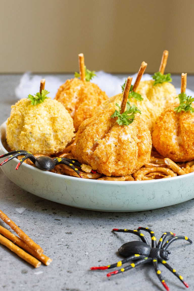 A plate of round, breaded cheese balls designed to look like pumpkins, topped with pretzel sticks and garnished with parsley, accompanied by plastic spider decorations.