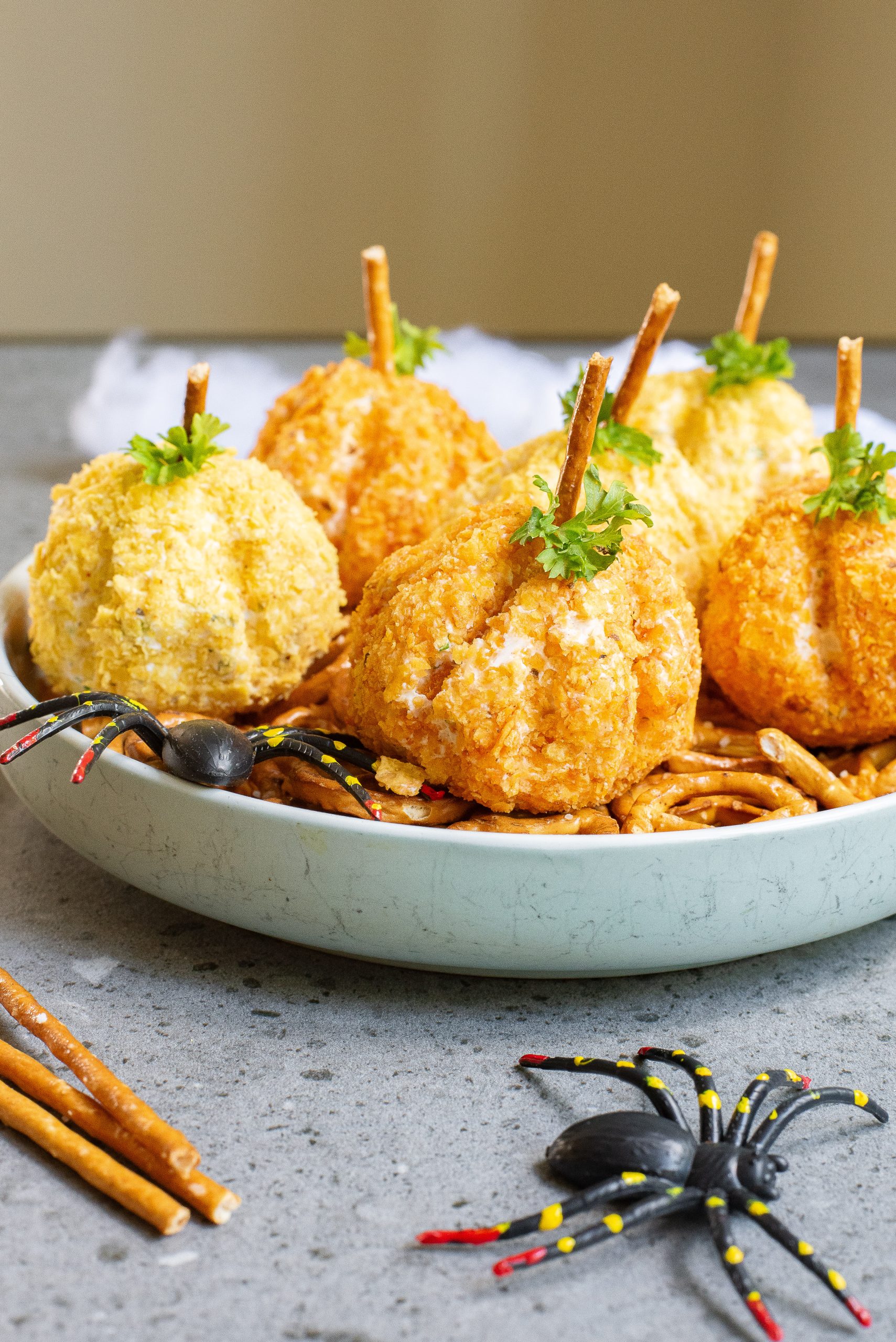 A plate of round, breaded cheese balls designed to look like pumpkins, topped with pretzel sticks and garnished with parsley, accompanied by plastic spider decorations.