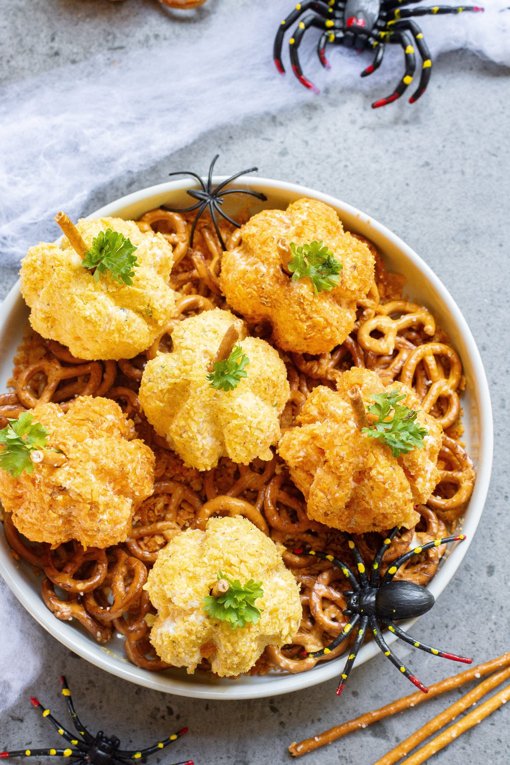 A plate of pumpkin-shaped cheese balls with pretzel stems is placed on a bed of pretzels, garnished with parsley. Decorative plastic spiders are arranged around the plate.