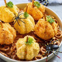 A plate of pumpkin-shaped cheese balls with pretzel stems, garnished with herbs, surrounded by Halloween-themed plastic spiders.