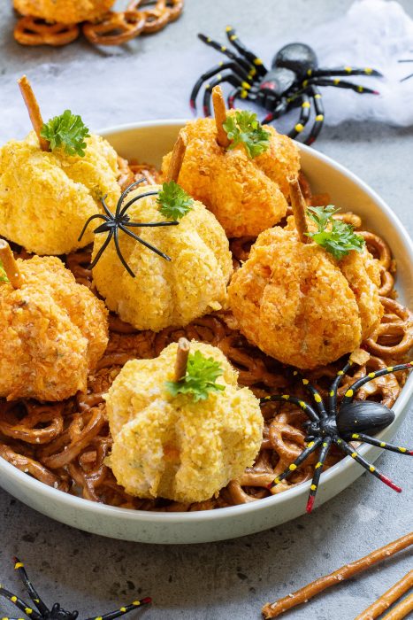 A plate of pumpkin-shaped cheese balls with pretzel stems, garnished with herbs, surrounded by Halloween-themed plastic spiders.