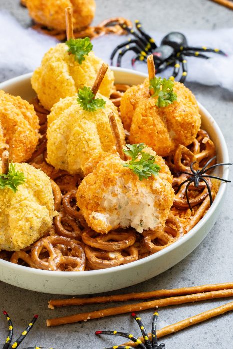 A bowl filled with pretzels and cheese balls shaped like pumpkins, topped with sticks to resemble stems and garnished with parsley. Plastic spider decorations are placed around the bowl.