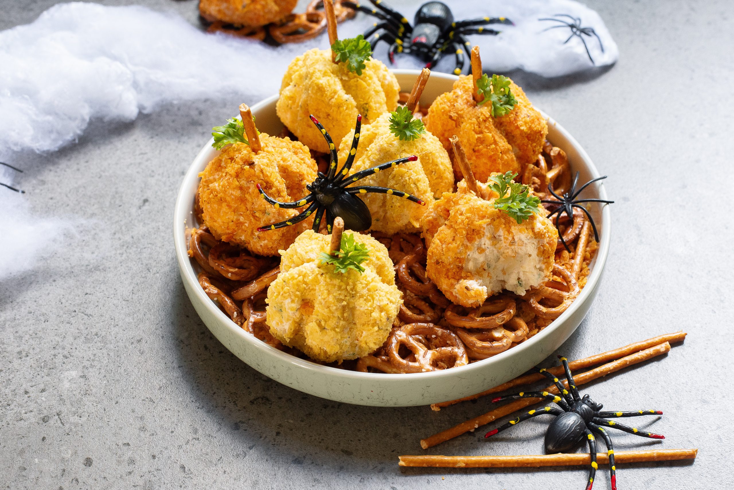 A bowl of pretzels topped with cheese balls decorated to look like pumpkins, garnished with parsley and surrounded by fake spiders and cobwebs on a gray surface.