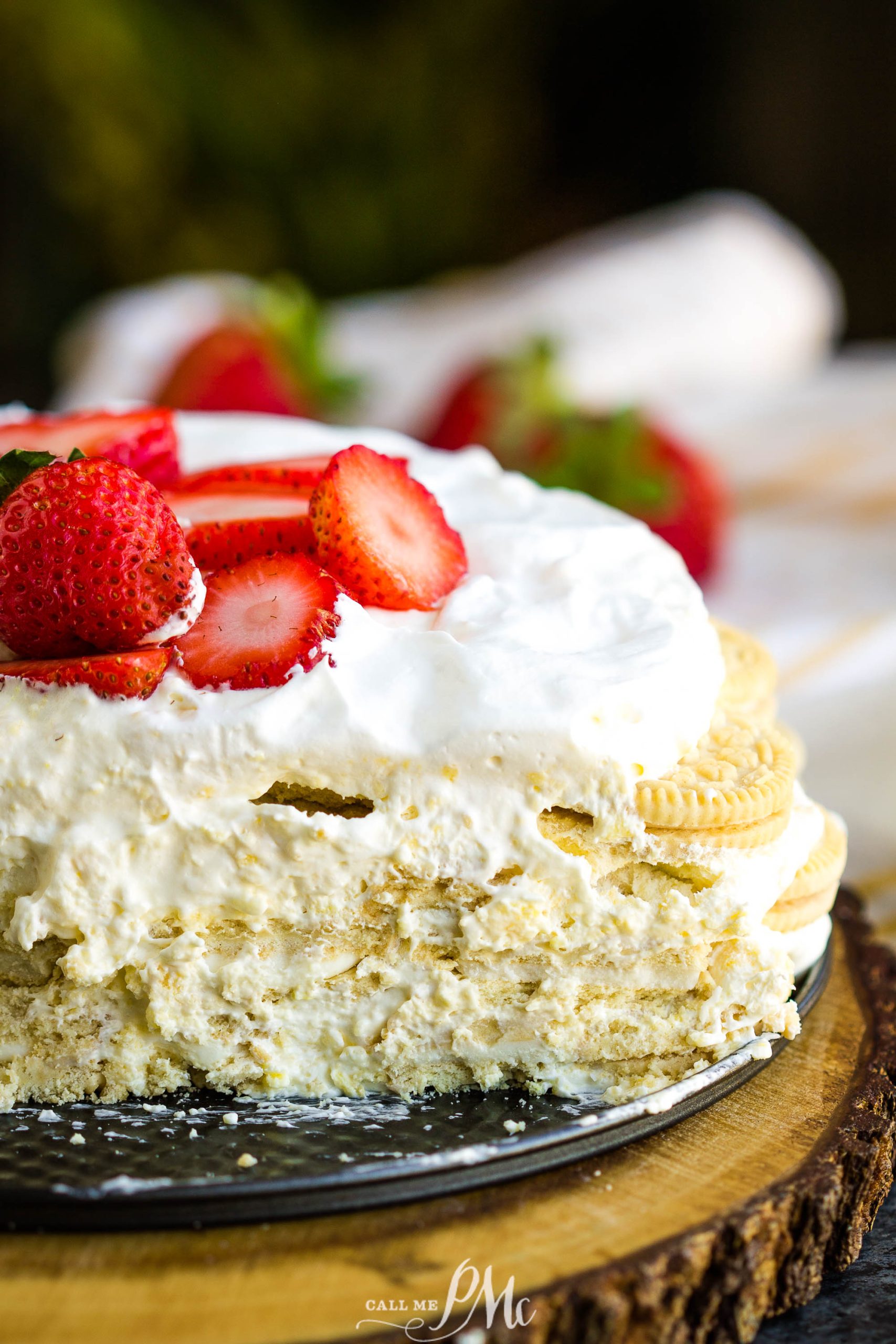 Close-up of a layered key lime icebox cake dessert with white cream, crushed cookies, and sliced strawberries on top, displayed on a wooden surface.