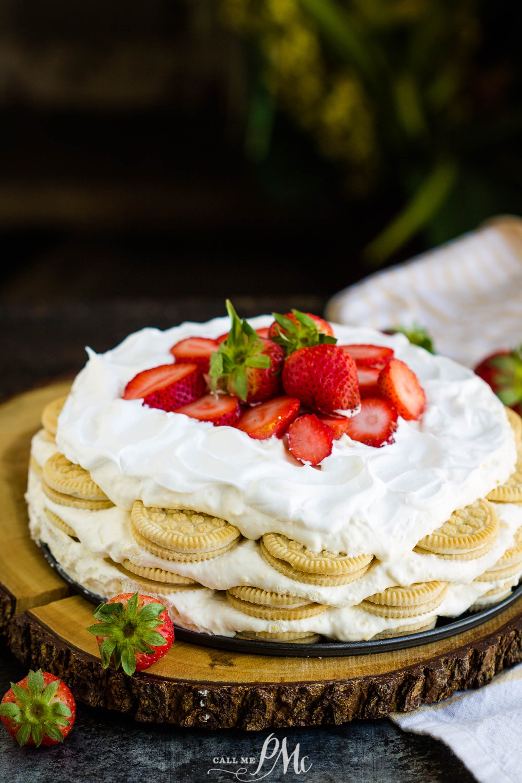 A dessert featuring layers of vanilla sandwich cookies, whipped cream, and sliced strawberries on top, displayed on a wooden board.