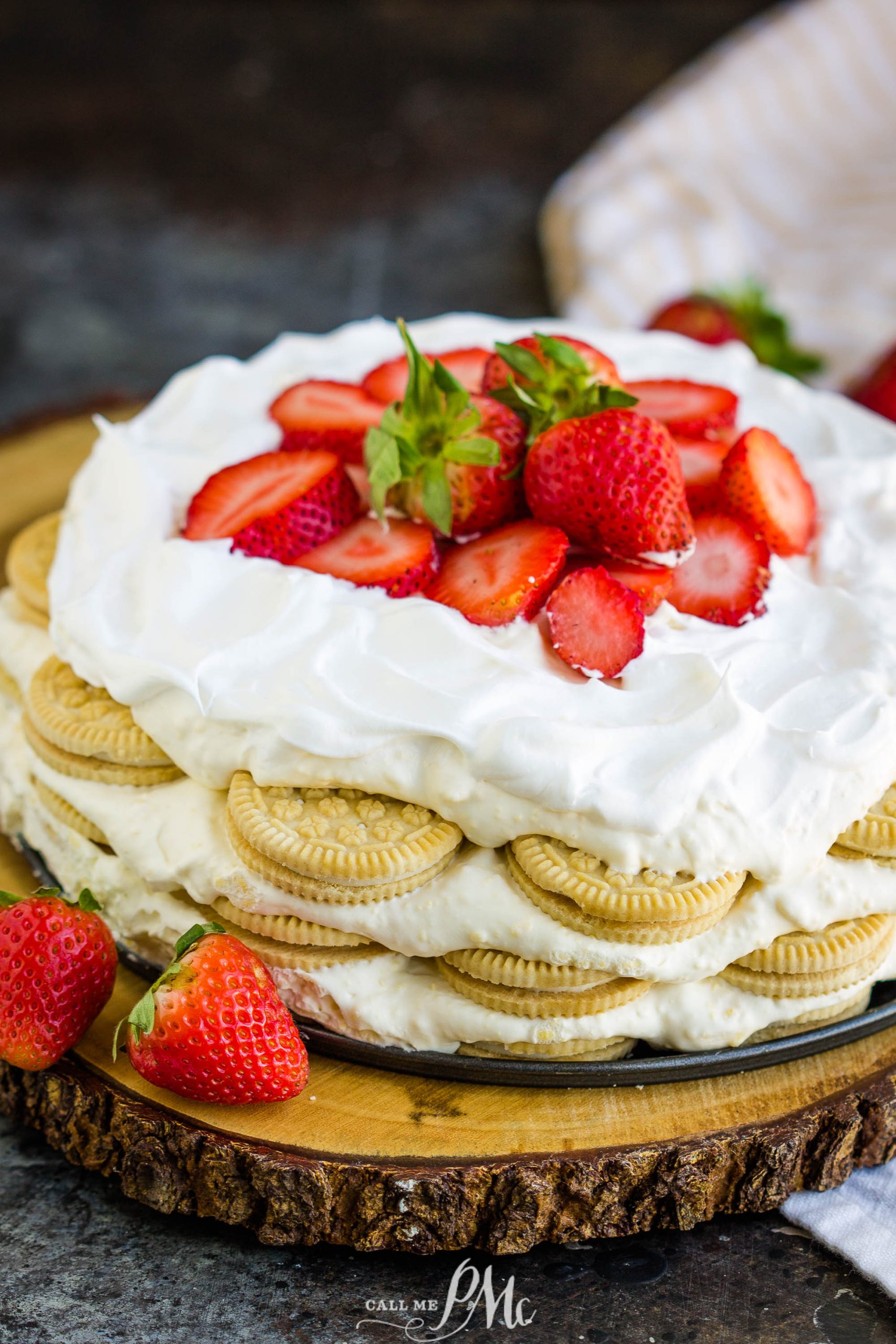 A no-bake cake made with layers of cookies, whipped cream, and topped with fresh strawberries is displayed on a wooden board.