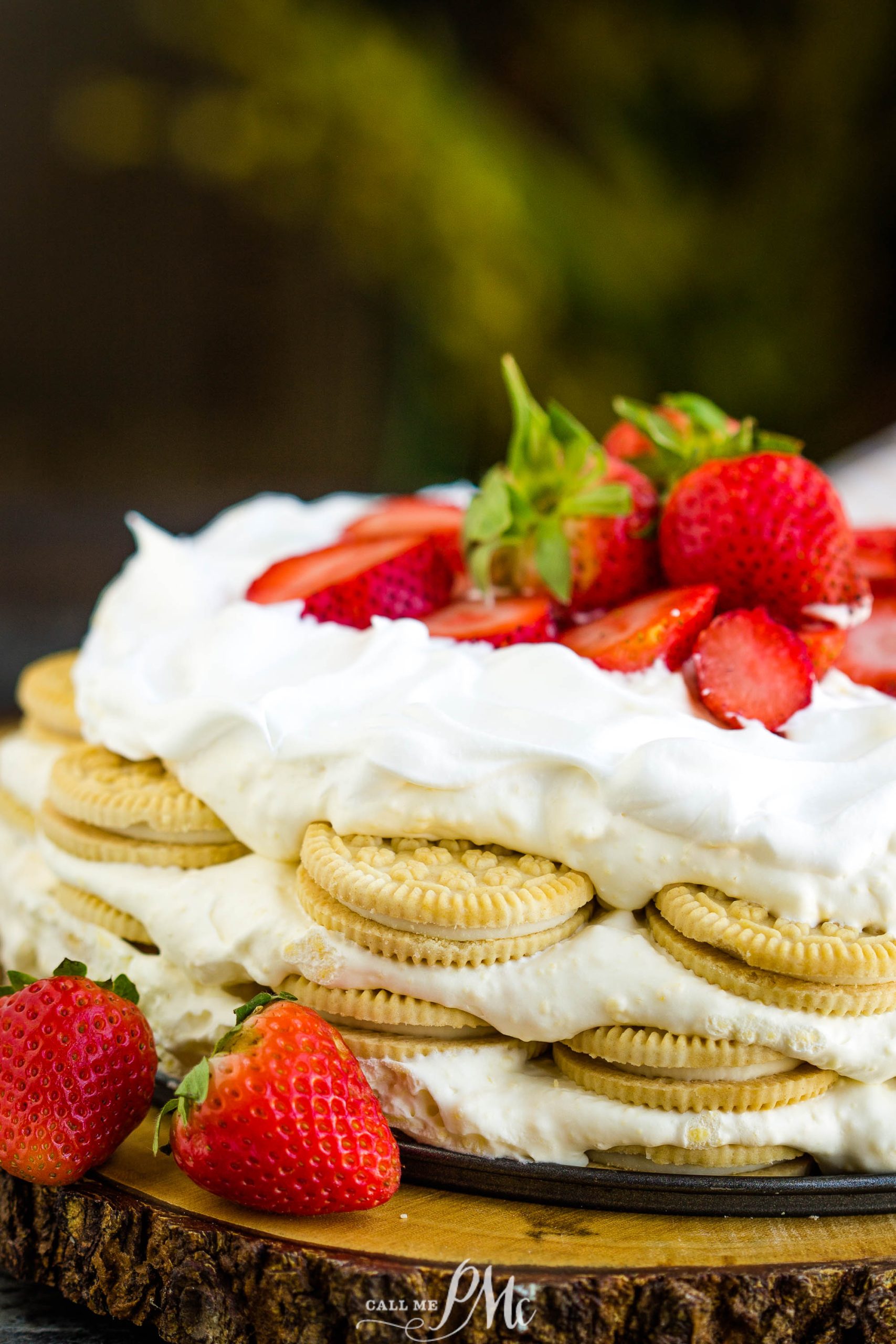 A layered dessert with cookies and whipped cream topped with fresh strawberries, displayed on a wooden surface with additional strawberries on the side.