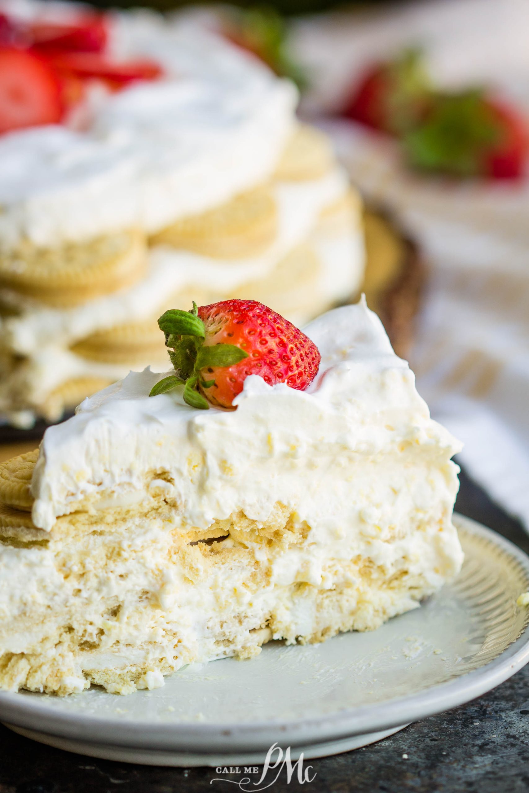 A slice of layered dessert with cream filling and topped with a strawberry on a white plate. The full dessert is visible in the background, also topped with strawberries.