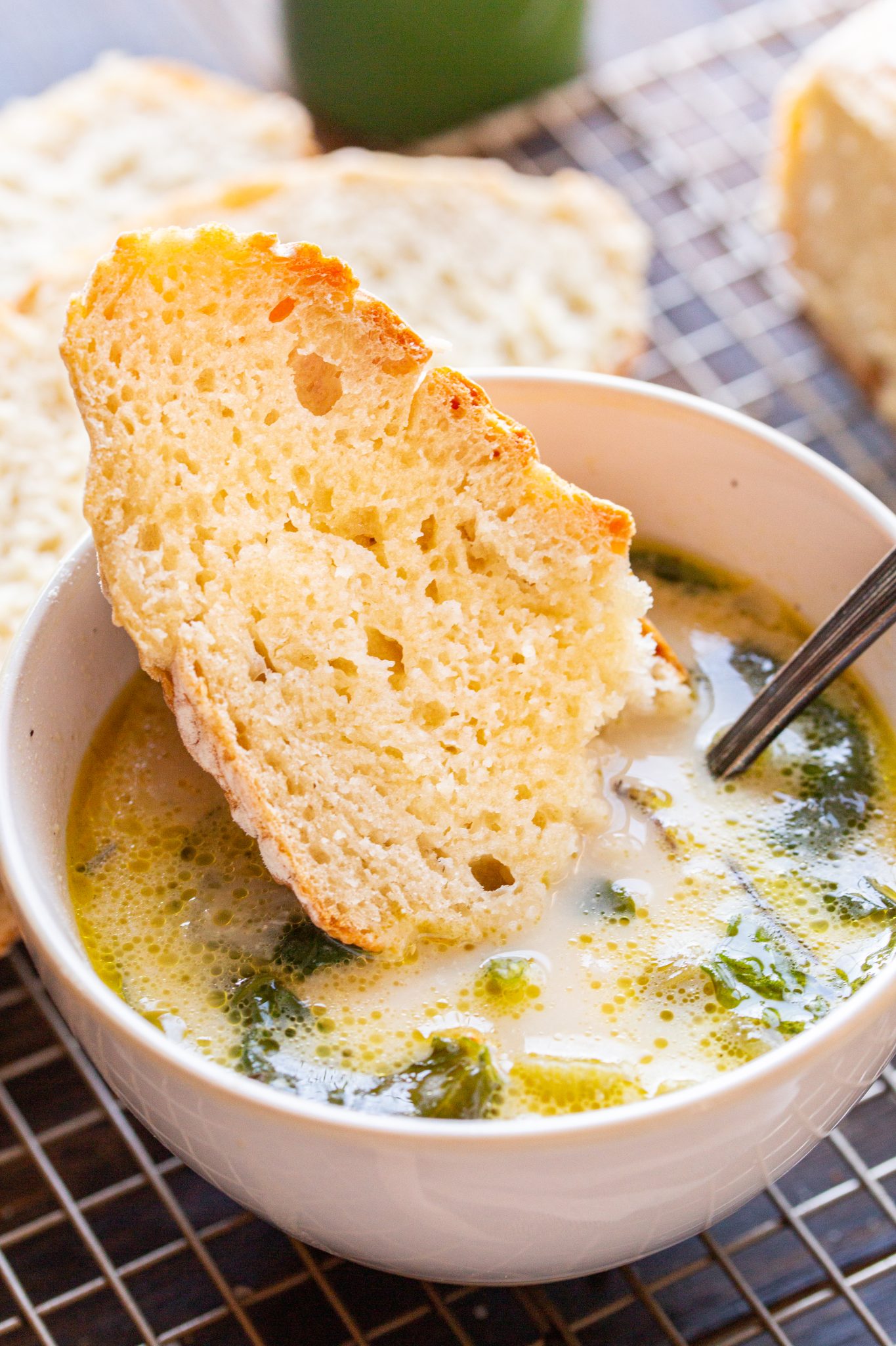 A bowl of soup with a piece of bread resting on the rim. A spoon is in the bowl, and there is bread in the background.