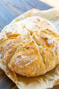 A round loaf of freshly baked bread sits on parchment paper on a wooden surface, with a golden, crispy crust and visible flour dusting.