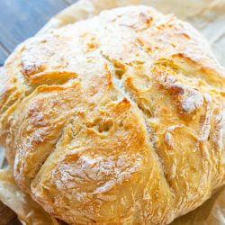 A round loaf of freshly baked bread sits on parchment paper on a wooden surface, with a golden, crispy crust and visible flour dusting.