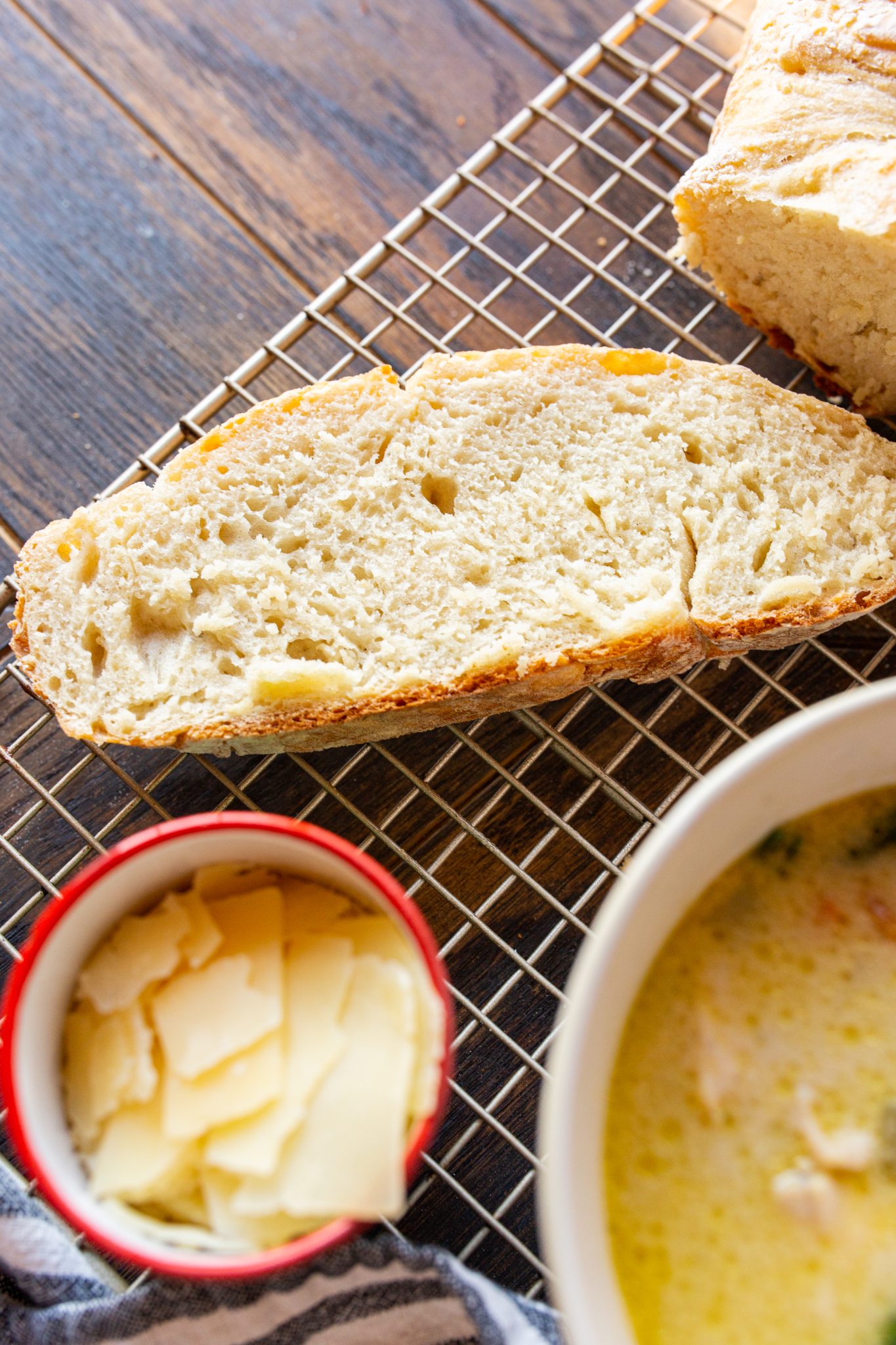 A slice of bread on a cooling rack beside a red bowl containing thin sliced cheese and a white dish with liquid contents on a wooden table.