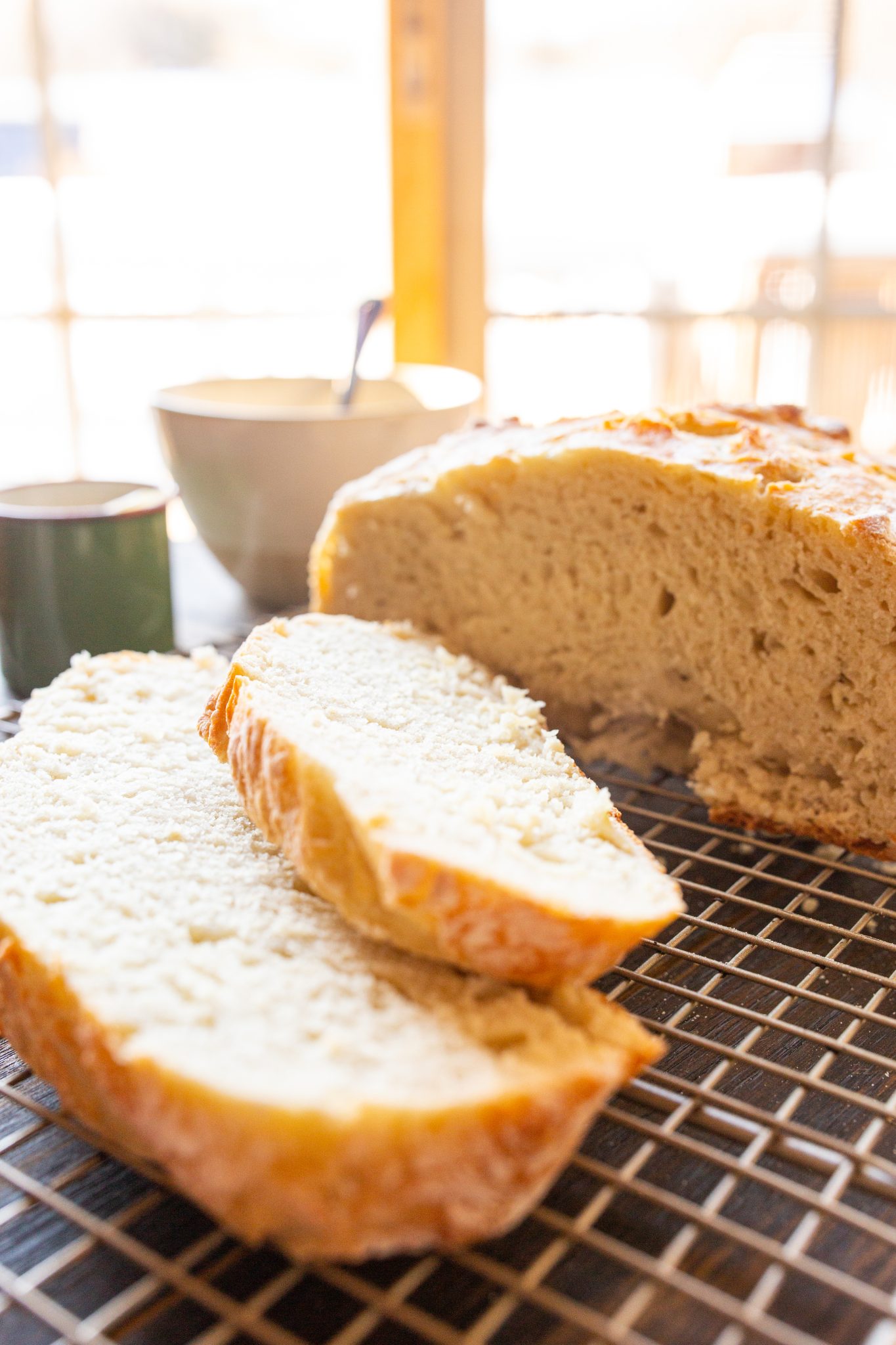 A loaf of Dutch Oven No Knead Bread on a cooling rack, partially sliced, with a bowl and two cups in the background.