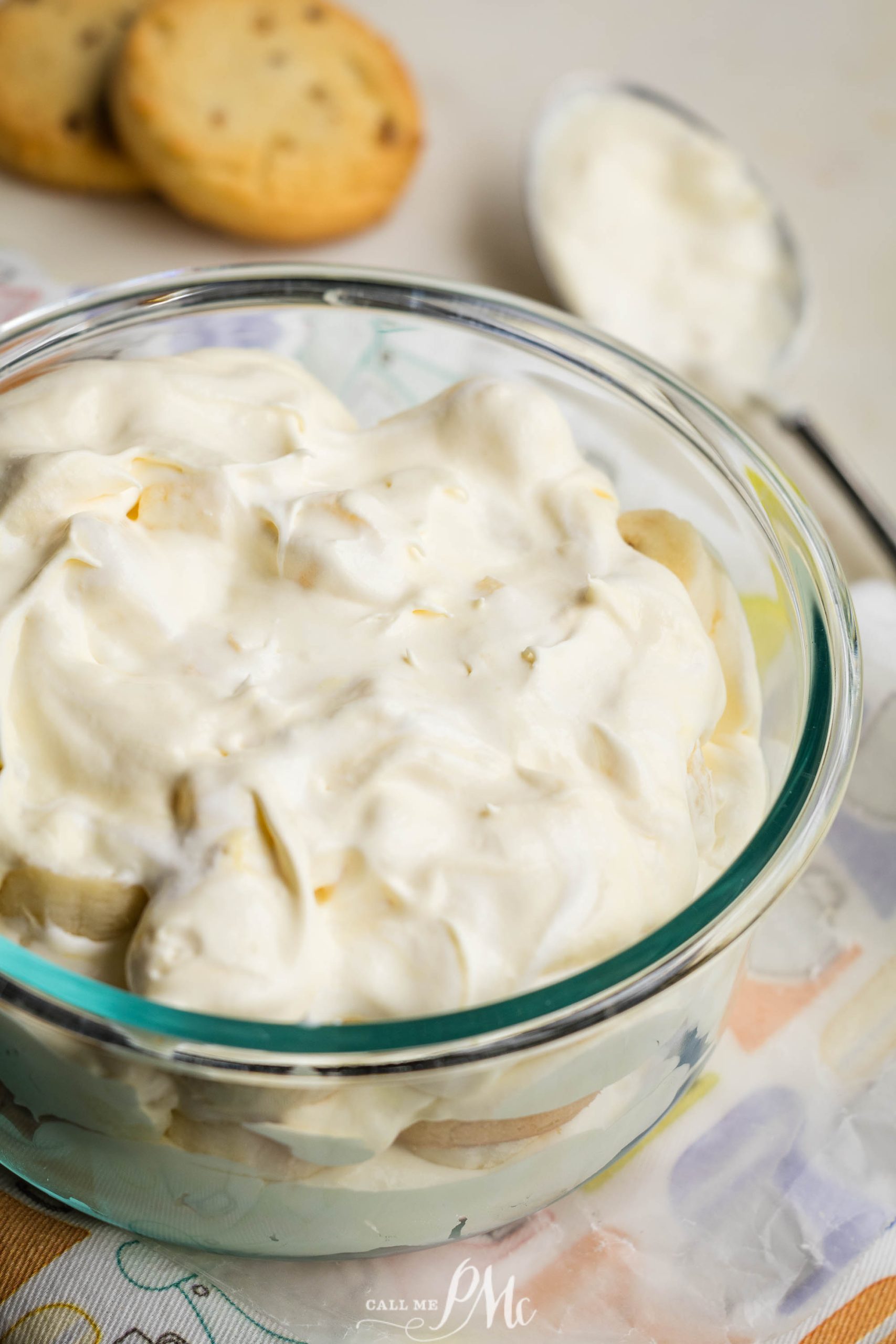 Glass bowl filled with a Pecan Sandies Banana Pudding dessert topped with whipped cream. Two cookies are partially visible in the background along with a spoon on a patterned cloth.