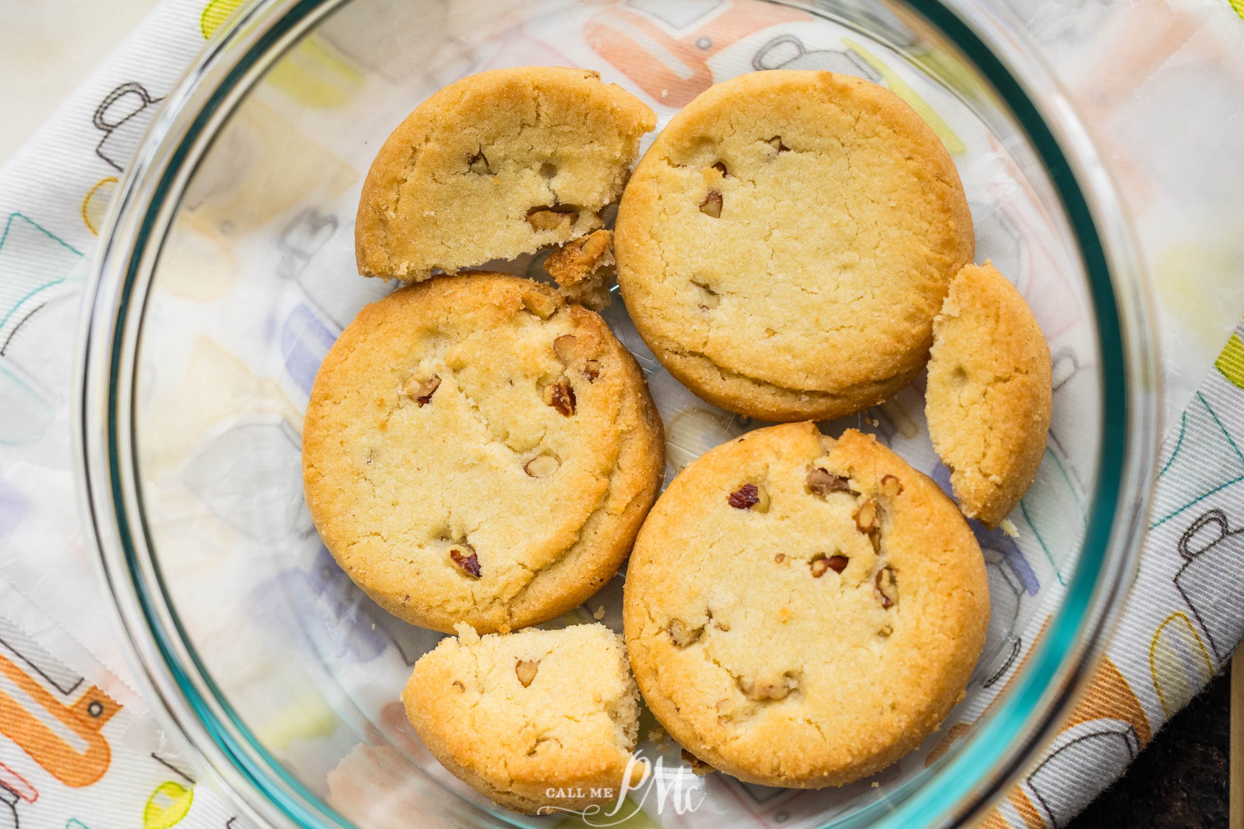 A glass bowl containing five chocolate chip cookies, with one cookie broken into pieces, placed on a white cloth with a colorful pattern.