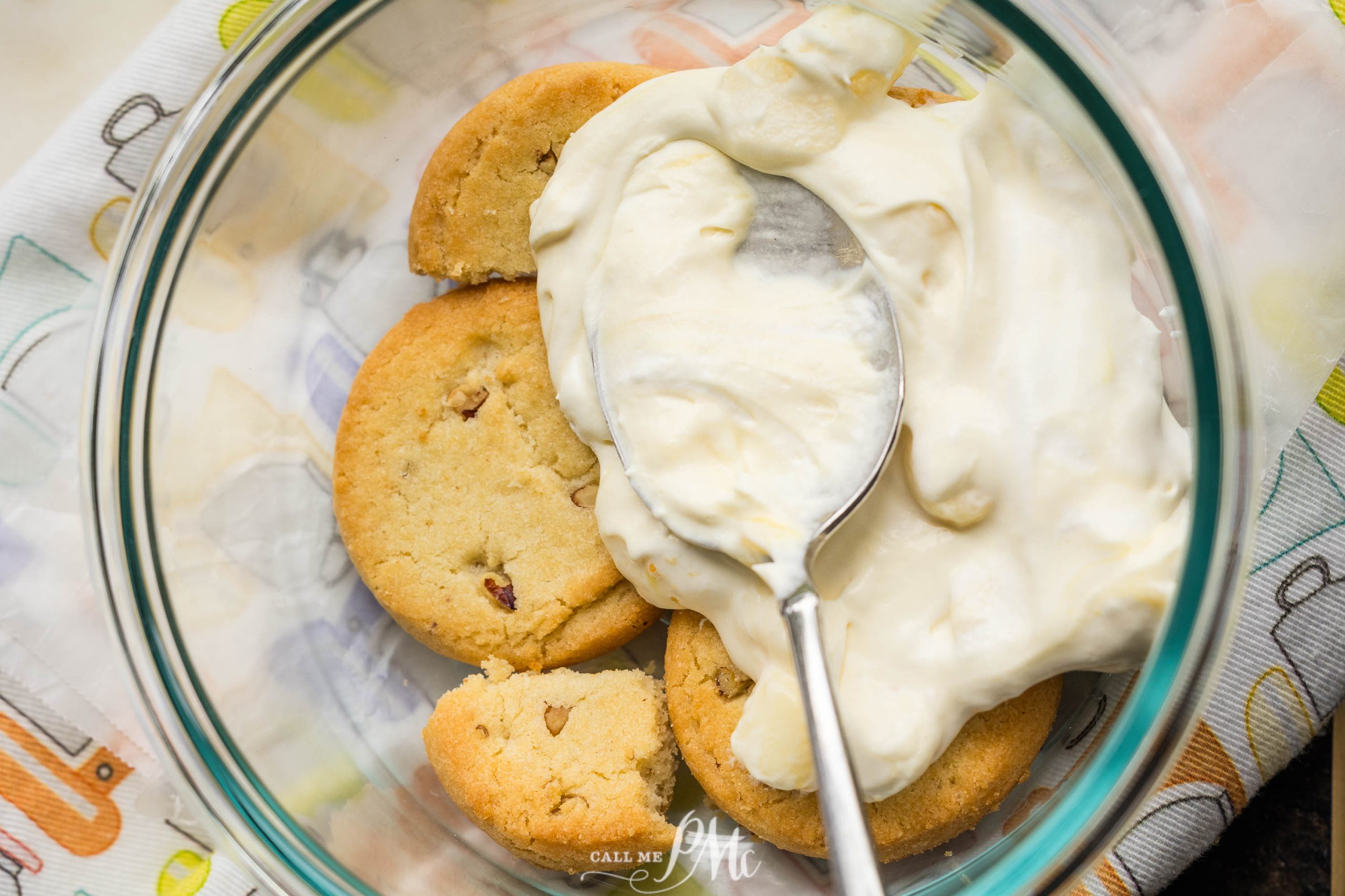 A glass bowl containing several cookies and a large spoon of creamy white mixture, likely frosting or cream.