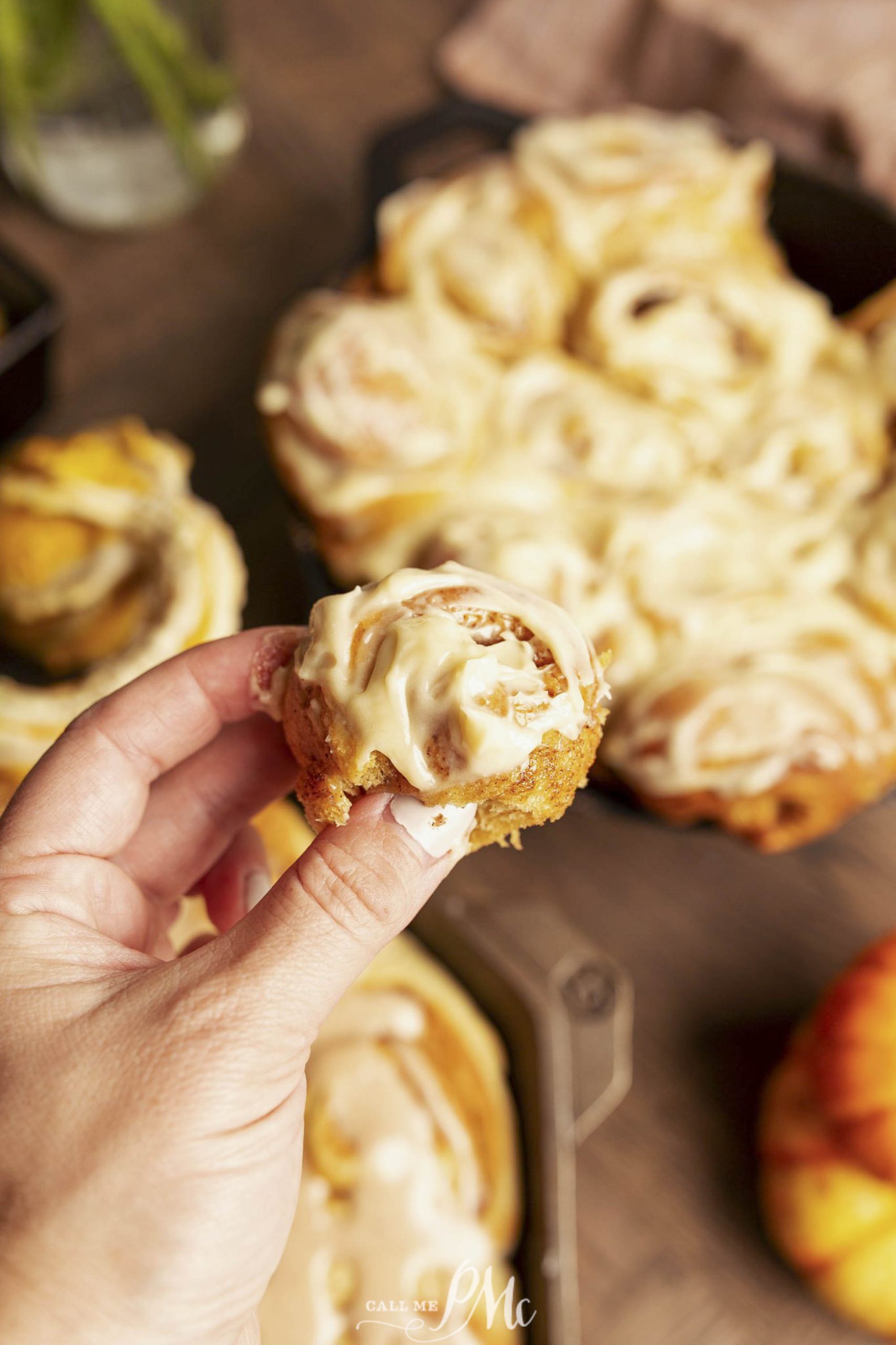 A hand holds a frosted cinnamon roll, with more cinnamon rolls visible in the background.