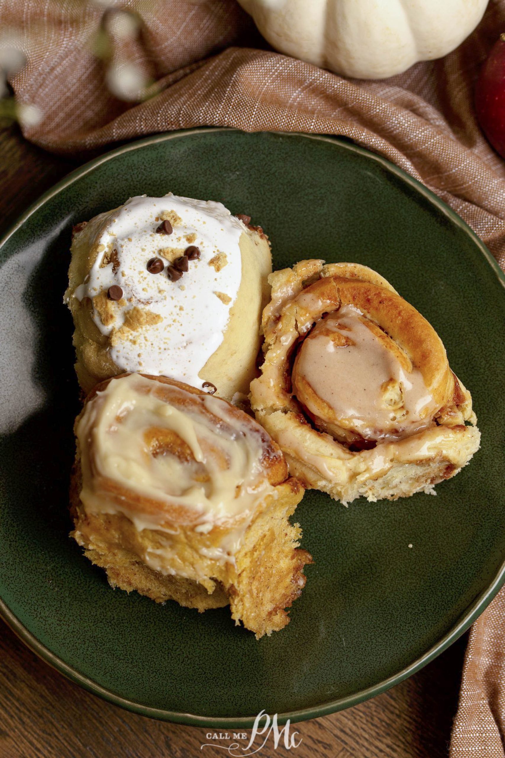 Three cinnamon rolls with different toppings are displayed on a green plate: one with frosting and sprinkles, one with icing, and one plain. A pumpkin and brown cloth are visible in the background.