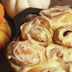 A close-up of freshly baked cinnamon rolls covered in icing, placed in a pan next to small white and orange pumpkins.