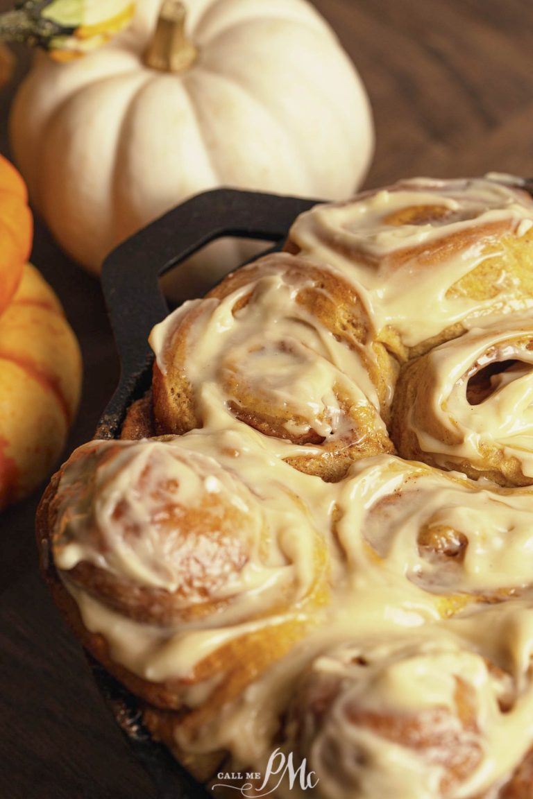 A close-up of freshly baked cinnamon rolls covered in icing, placed in a pan next to small white and orange pumpkins.