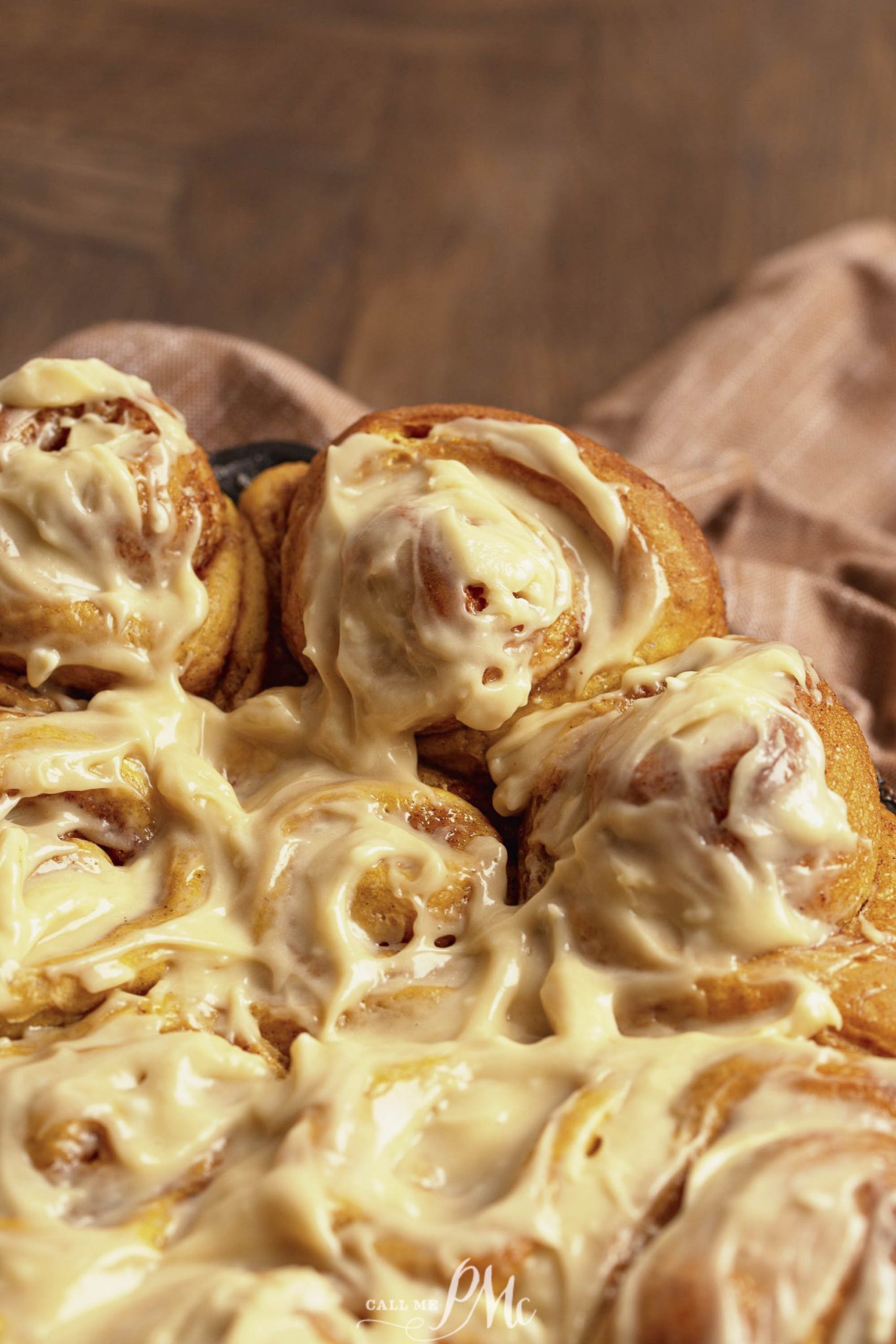 A close-up of freshly baked buns topped with a thick layer of creamy icing. The rolls are arranged closely together, showing a gooey and appetizing texture.