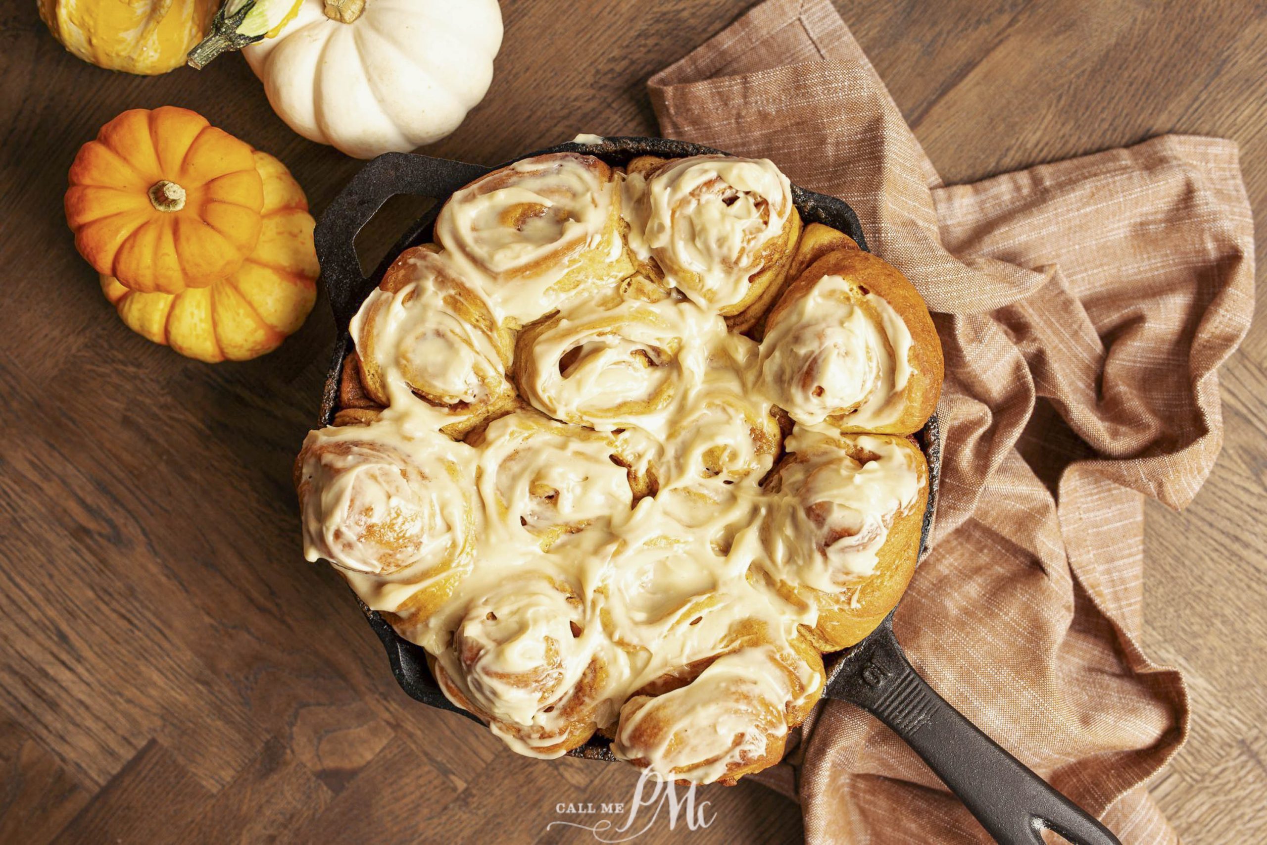 A skillet of frosted buns on a wooden table, surrounded by small pumpkins and a brown cloth.